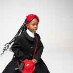 Young girl wearing red jeweled velvet bow headband with black coat and red apple bag