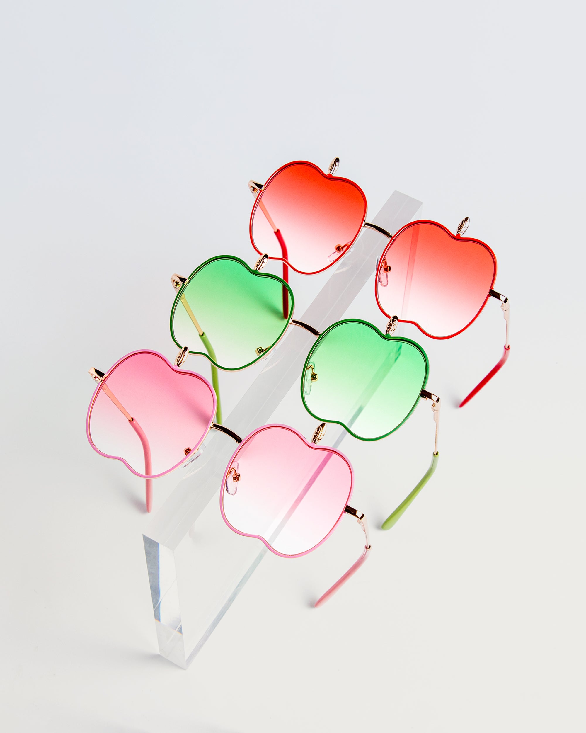 Five pairs of apple-shaped kids sunglasses in red, pink, and green on a clear acrylic stand