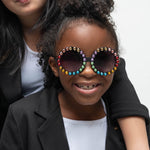 Girl smiling while wearing Carrousel Color sunglasses with rainbow rhinestones, dressed in a black blazer