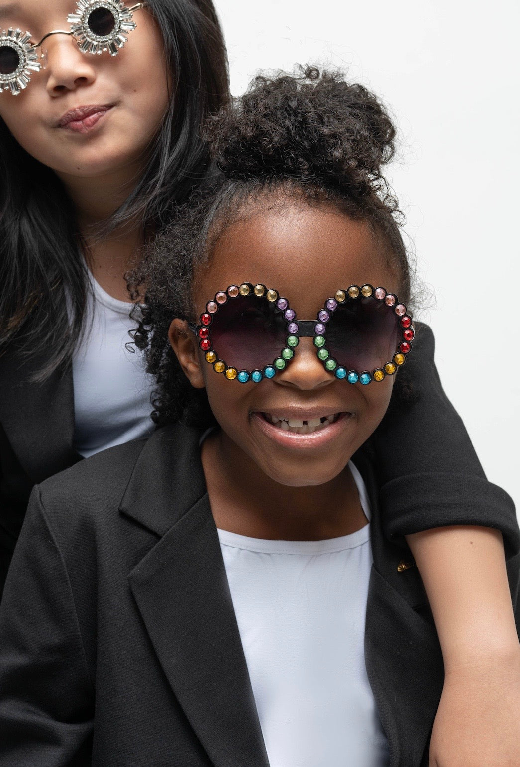 Girl smiling while wearing Carrousel Color sunglasses with rainbow rhinestones, dressed in a black blazer