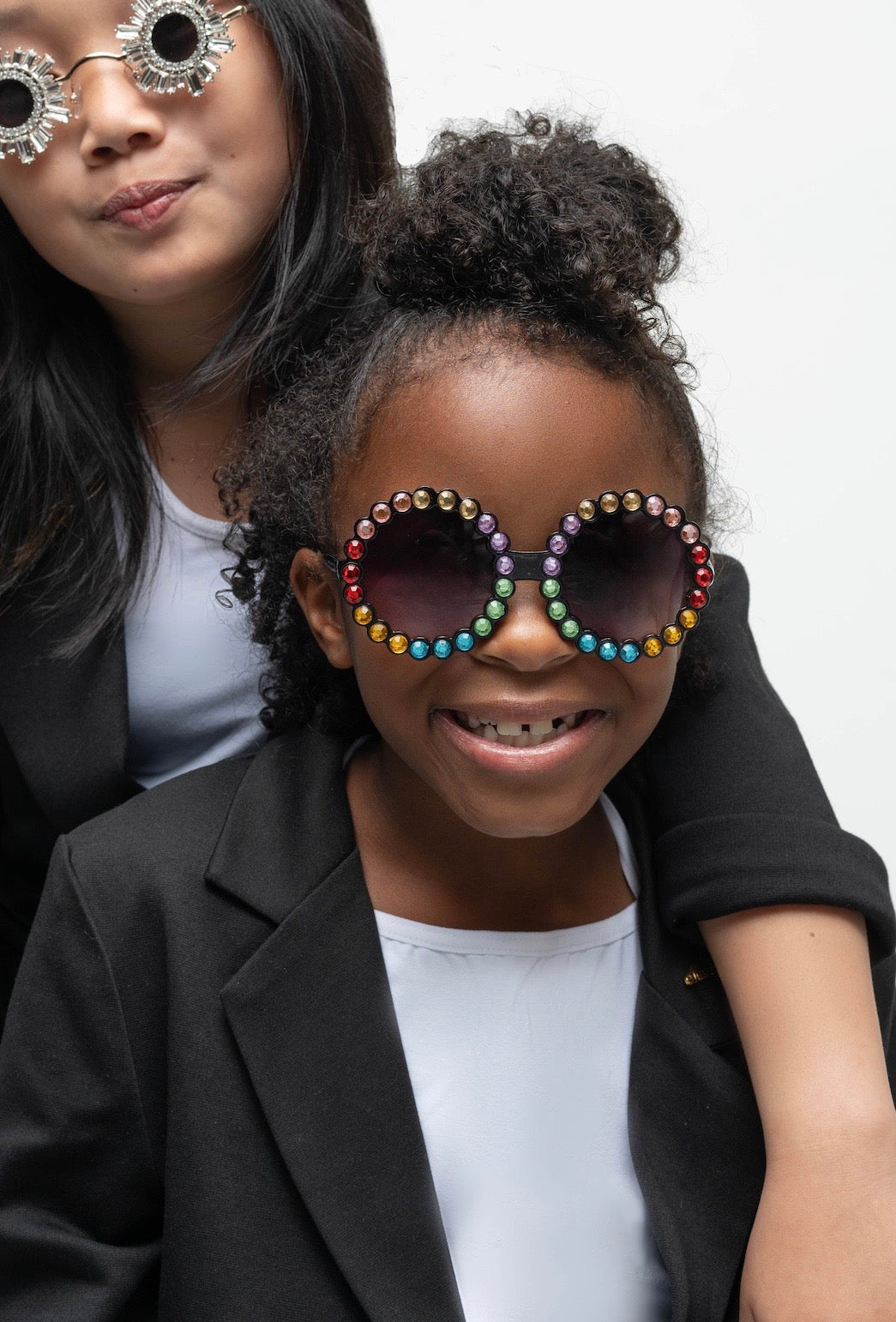 Girl smiling while wearing Carrousel Color sunglasses with rainbow rhinestones, dressed in a black blazer
