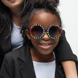 Girl smiling while wearing Carrousel Color sunglasses with rainbow rhinestones, dressed in a black blazer
