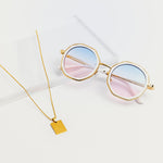 Goddess Cream sunglasses with pink and blue gradient lenses displayed next to gold necklace on white surface
