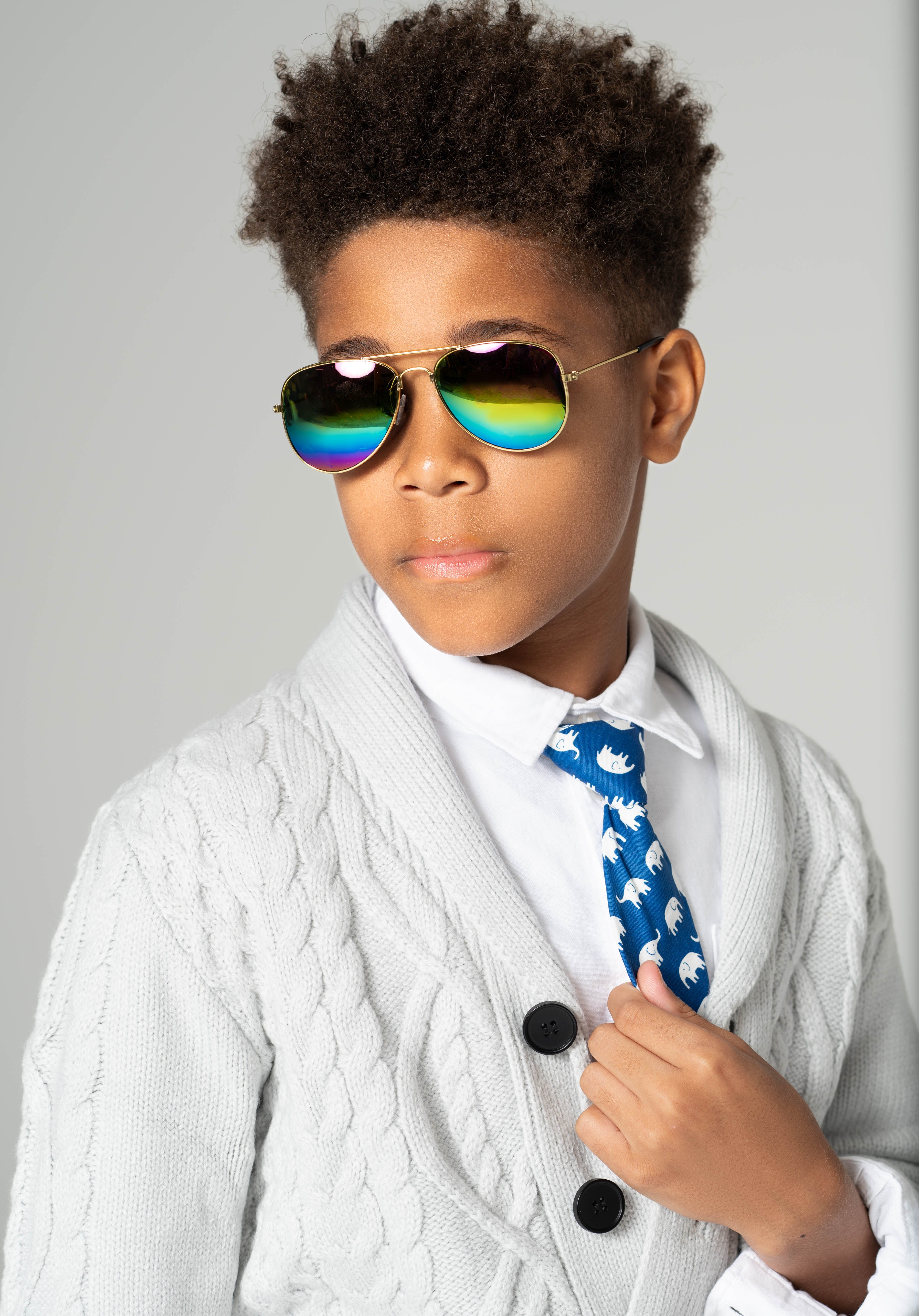 Boy wearing Rainbow Gleam sunglasses with gold frames and reflective rainbow lenses, styled in a white shirt and knit cardigan