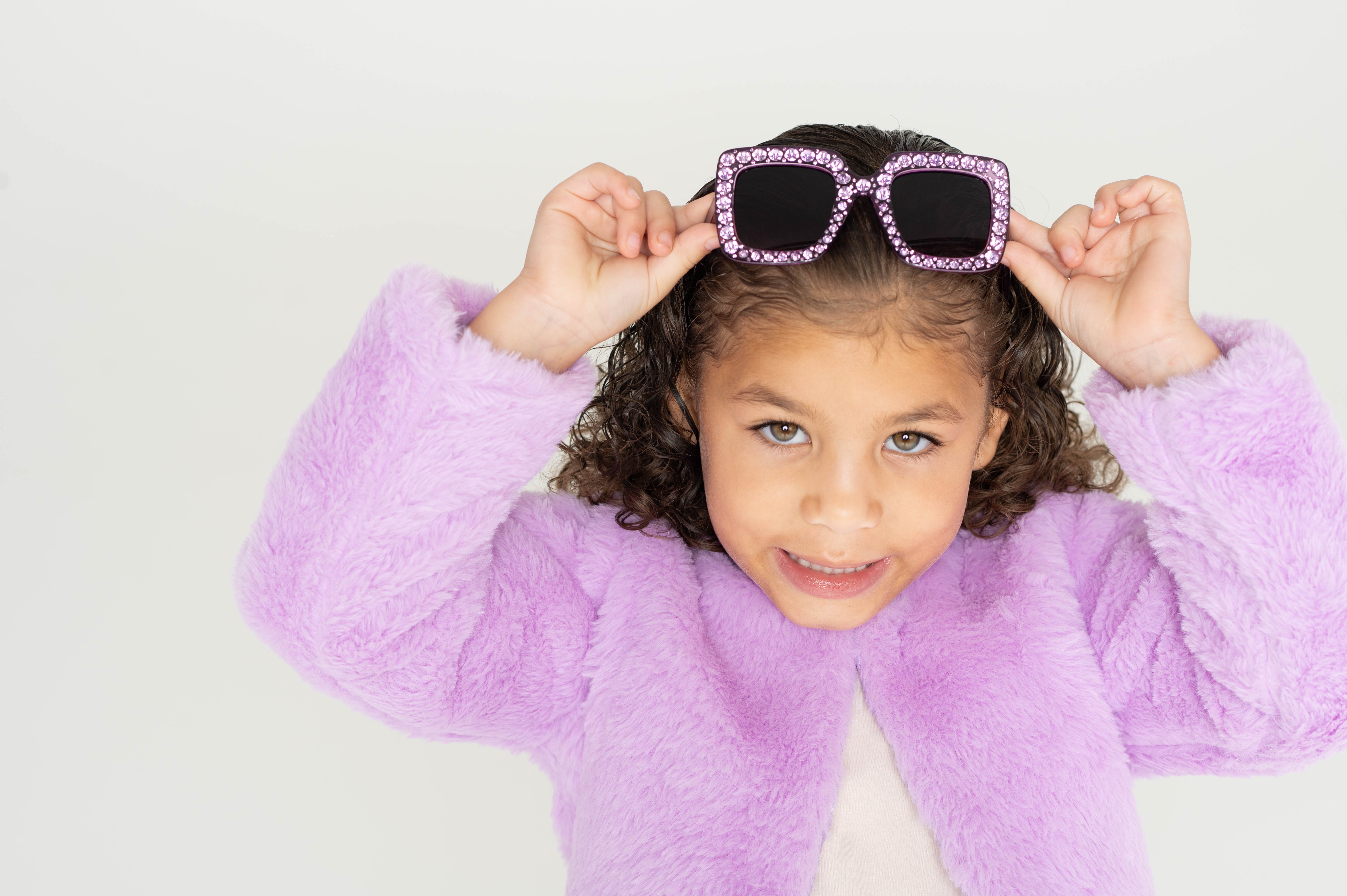 Girl smiling while lifting Sparkle Lilac Blooms sunglasses, wearing a lilac faux fur jacket